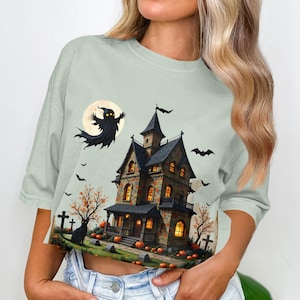 May include: A light green crop top featuring a Halloween-themed graphic. The design includes a haunted house, a ghost, bats, pumpkins, and a full moon. The artwork is detailed with a spooky and festive atmosphere.