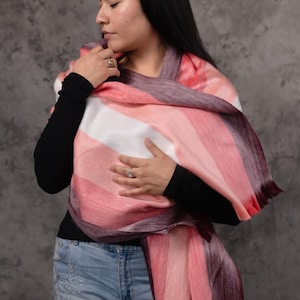 May include: A colourful scarf with a striped pattern in shades of pink, white, and burgundy. The scarf is draped over the shoulders. The scarf has a fringed edge. The scarf is made of a soft material.