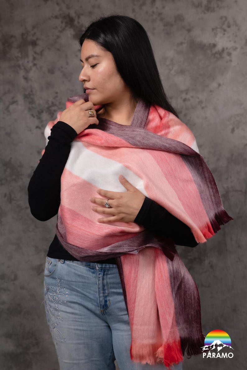 May include: A colourful scarf with a striped pattern in shades of pink, white, and burgundy. The scarf is draped over the shoulders. The scarf has a fringed edge. The scarf is made of a soft material.