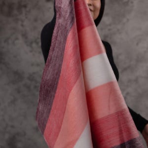 May include: A striped blanket in shades of red, pink, and white, with a dark purple border. The blanket is held up, displaying its soft texture and design. The blanket is made of a soft material.