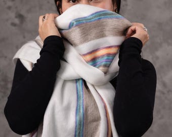 May include: A white scarf with colourful stripes of blue, green, and brown is held up to the face. The scarf is wrapped around the neck and shoulders. The background is a solid grey colour. The scarf has a tag on the bottom left corner.