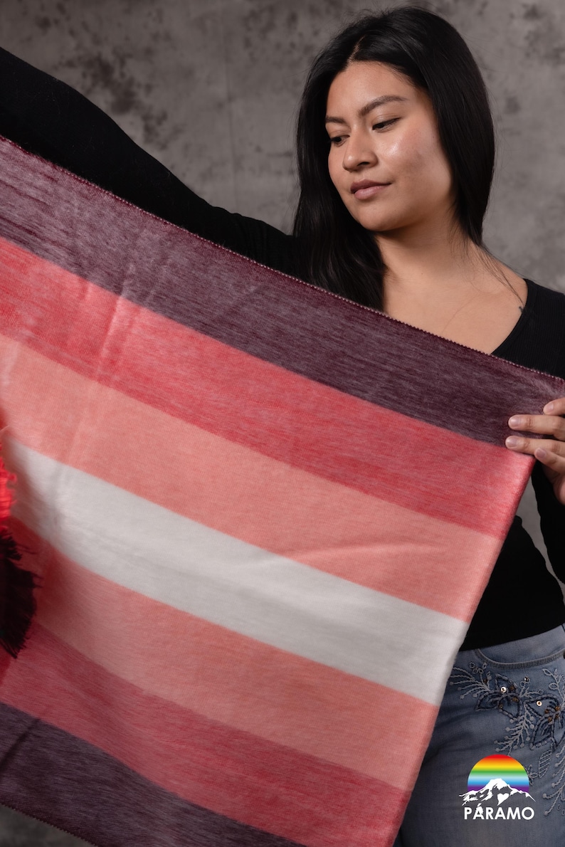 May include: A rectangular scarf with horizontal stripes in shades of red, pink, white, and purple. The scarf is held up by a person, showcasing the soft texture and colour variations. The scarf is a fashion accessory.