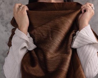 May include: A brown scarf with a soft, textured appearance. The scarf is held up, partially obscuring the face of a person wearing a white shirt. The scarf's colour ranges from light to dark brown. The background is a mottled grey.