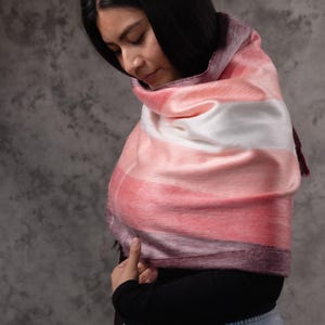 May include: A soft, striped scarf in shades of pink, white, and purple. The scarf is draped over the shoulders. The scarf has a silky texture and a dark purple fringe.