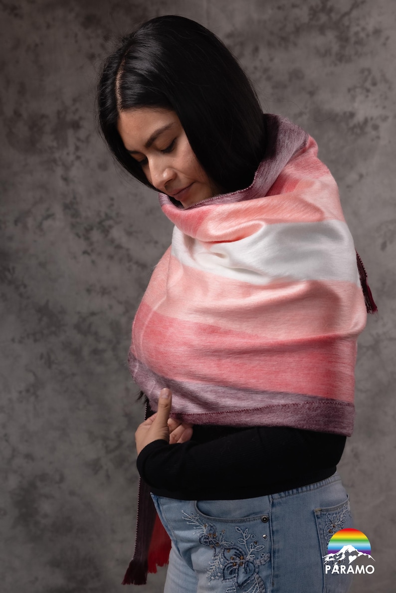 May include: A soft, striped scarf in shades of pink, white, and purple. The scarf is draped over the shoulders. The scarf has a silky texture and a dark purple fringe.