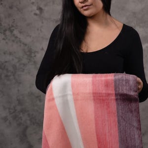 May include: A striped blanket in shades of pink, white, and purple. The blanket is held by a person wearing a black top. The blanket has a logo in the bottom right corner.