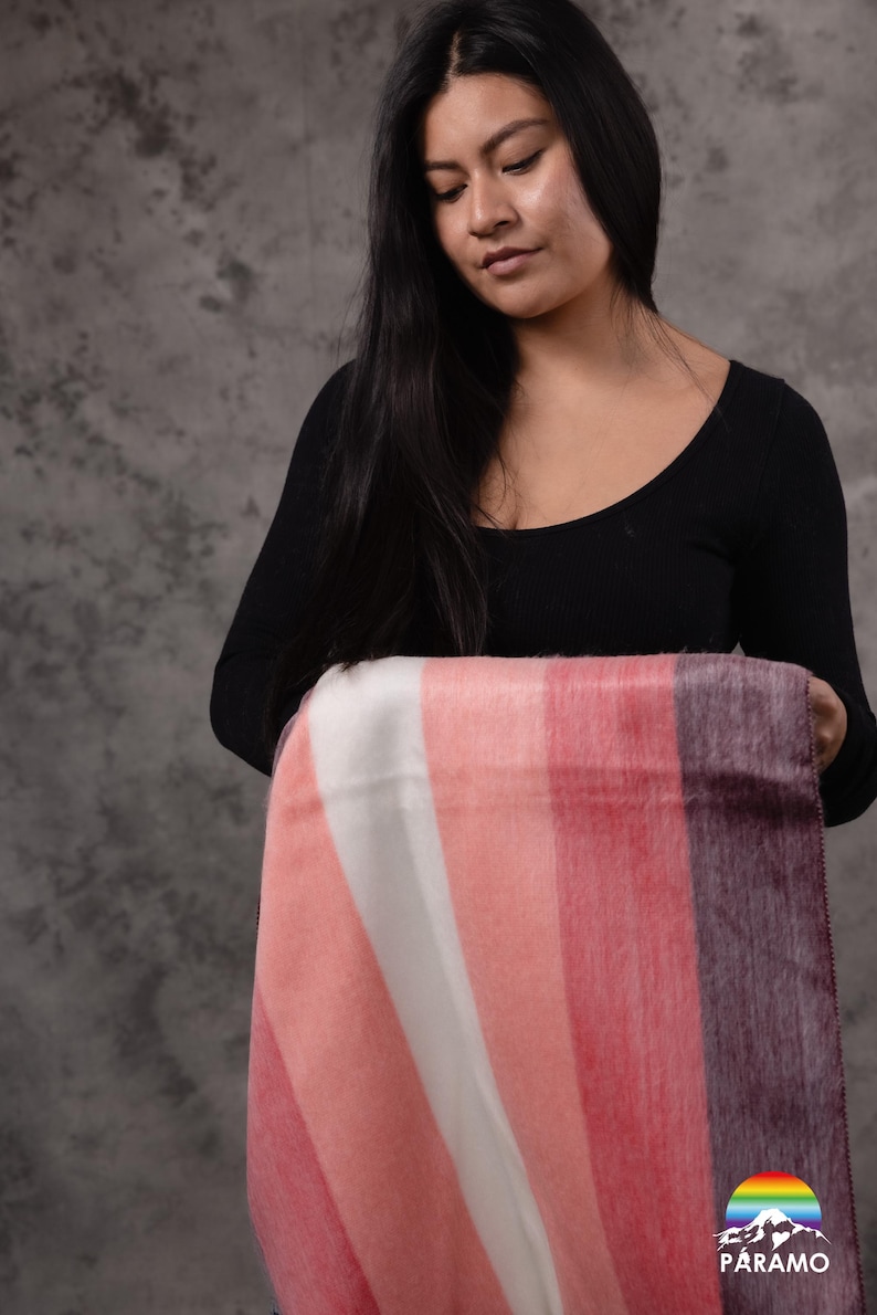 May include: A striped blanket in shades of pink, white, and purple. The blanket is held by a person wearing a black top. The blanket has a logo in the bottom right corner.