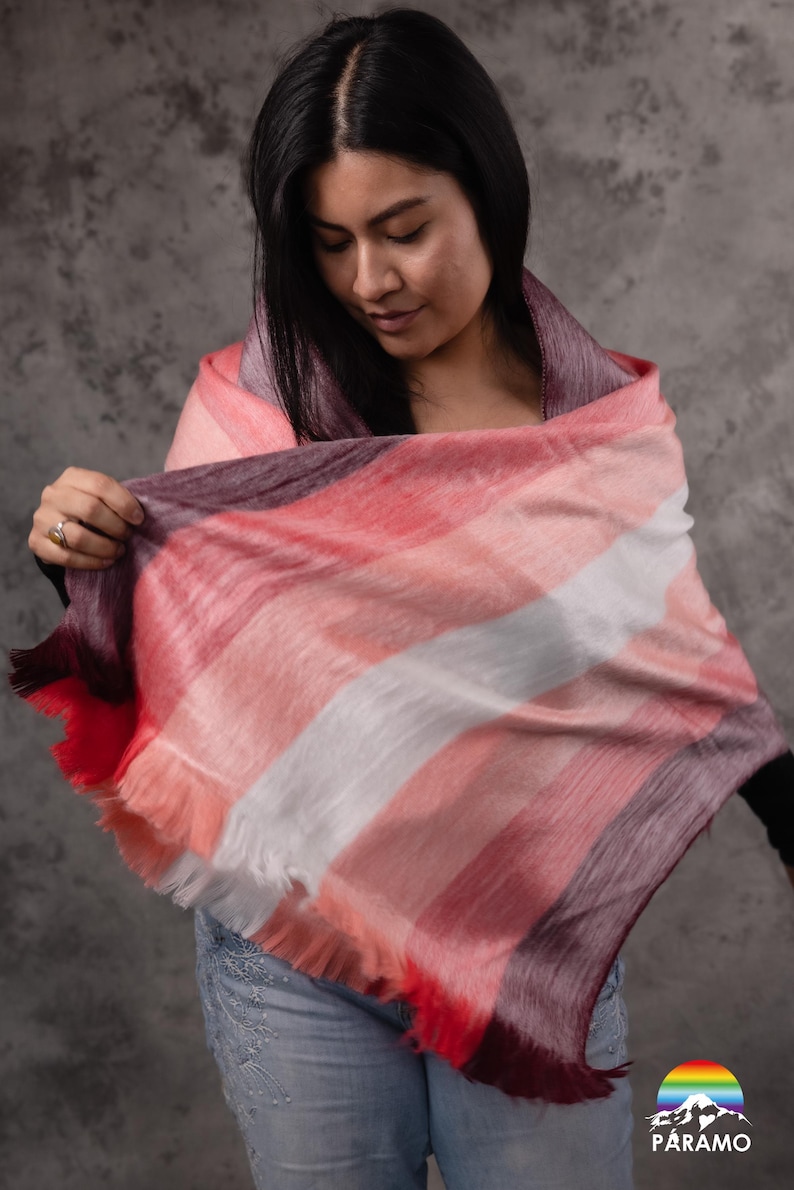 May include: A patterned scarf in red, pink, white, and purple stripes, with a fringed edge. The scarf is draped over the shoulders. The Paramo logo is visible in the bottom right corner. The scarf is a stylish accessory.