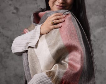 May include: A striped shawl in shades of pink, grey, white, and brown, with a fringed edge. The model is wearing a cream-coloured sweater. The Paramo logo is visible in the bottom right corner. The shawl is a cosy accessory.