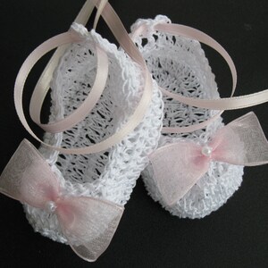 May include: A pair of white crocheted baby booties with pink ribbon ties and sheer bows. Each bow has a small pearl bead in the center. The booties are a delicate handmade item, perfect for a newborn.