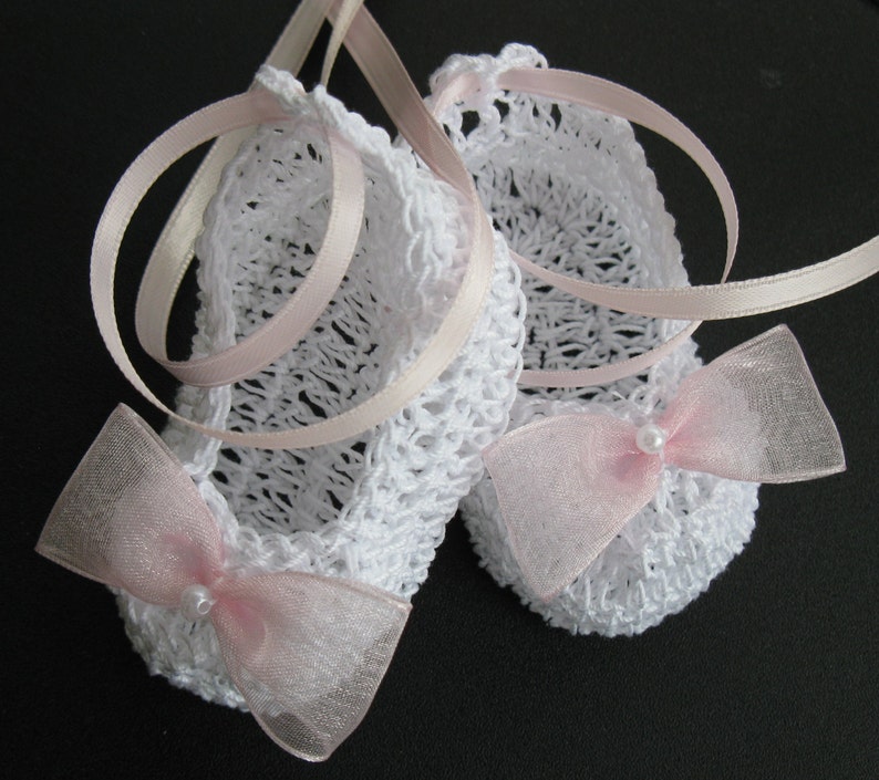 May include: A pair of white crocheted baby booties with pink ribbon ties and sheer bows. Each bow has a small pearl bead in the center. The booties are a delicate handmade item, perfect for a newborn.