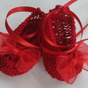 K&ouml;nnte beinhalten: Ein Paar handgefertigte rote Babyschuhe. Die Schuhe sind mit einem Lochmuster geh&auml;kelt und haben eine transparente rote Schleife auf der Spitze. Rote Satinb&auml;nder sind zum Binden angebracht.