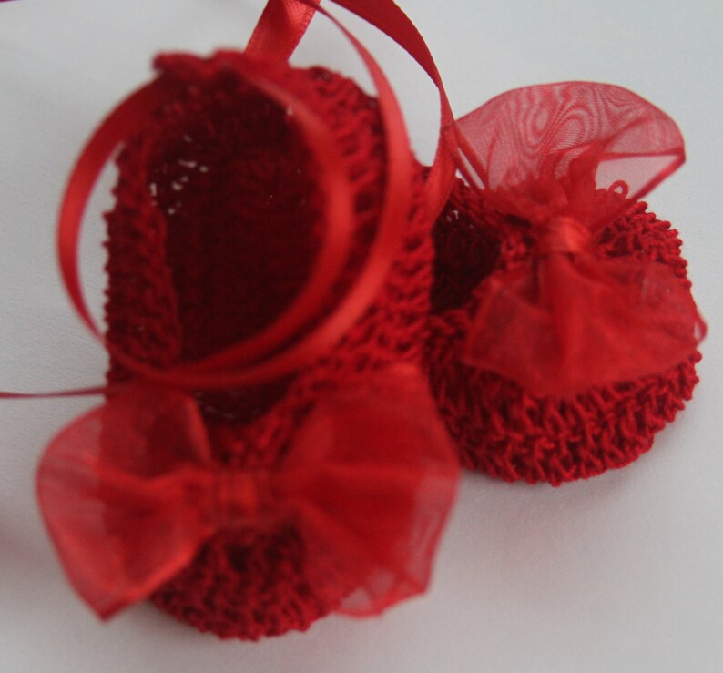 K&ouml;nnte beinhalten: Ein Paar handgefertigte, rote geh&auml;kelte Babyschuhe mit Satinbandschn&uuml;ren und transparenten Schleifen. Die Schuhe haben eine leuchtend rote Farbe und werden auf einer wei&szlig;en Oberfl&auml;che pr&auml;sentiert. Die Schleifen verleihen den handgefertigten Babyschuhen eine zarte Note.