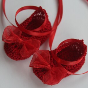 K&ouml;nnte beinhalten: Ein Paar kleine, rote geh&auml;kelte Babyschuhe mit roten Organza-Schleifen und passendem roten Band. Die Schuhe sind handgefertigt und haben ein zartes, offenes Design, perfekt f&uuml;r ein Neugeborenes oder Kleinkind. Das Band sorgt f&uuml;r eine dekorative Note.