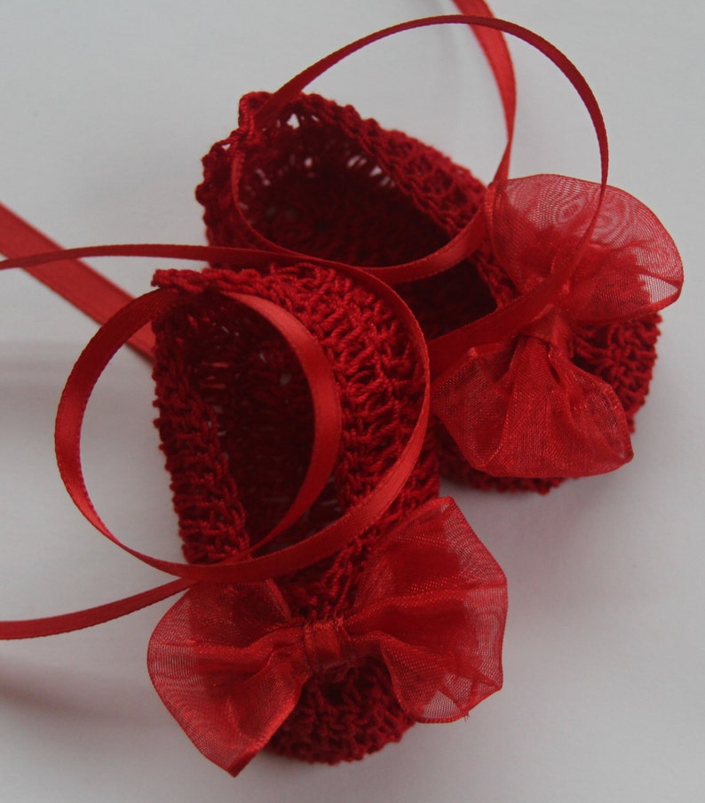 K&ouml;nnte beinhalten: Ein Paar kleine, rote geh&auml;kelte Babyschuhe mit zarten Schleifen und Satinb&auml;ndern. Die Schuhe sind leuchtend rot und haben ein strukturiertes Strickmuster. Die Schleifen sind aus einem transparenten, roten Stoff gefertigt.