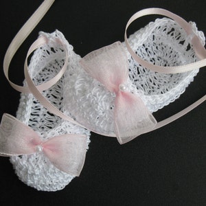 May include: A pair of white crocheted baby booties with pink bows and pearl accents. The booties are tied with a light pink ribbon. The open weave design is visible. The booties are a baby accessory.