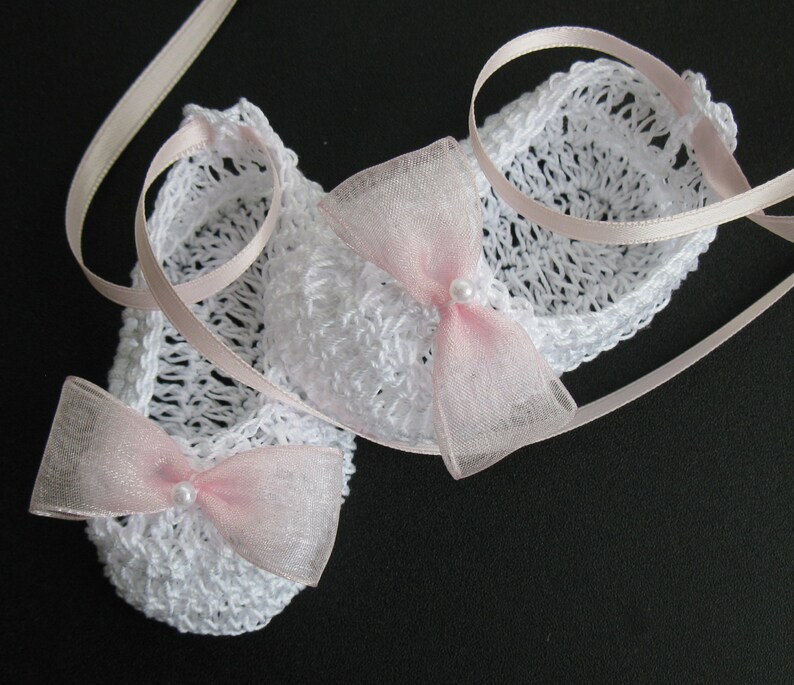 May include: A pair of white crocheted baby booties with pink bows and pearl accents. The booties are tied with a light pink ribbon. The open weave design is visible. The booties are a baby accessory.