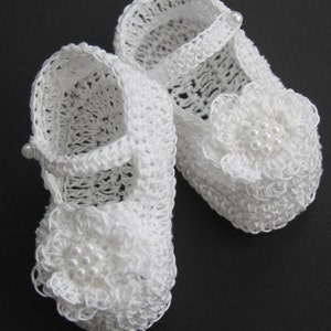 Peut inclure: Une paire de chaussons de b&eacute;b&eacute; blancs, crochet&eacute;s. Chaque chaussure pr&eacute;sente un ornement floral d&eacute;licat avec des perles et une bride avec un bouton perl&eacute;. Les chaussures sont con&ccedil;ues avec un motif ajour&eacute;, sugg&eacute;rant une construction l&eacute;g&egrave;re et a&eacute;r&eacute;e.