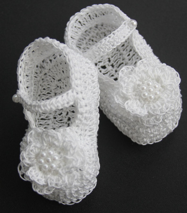 Peut inclure: Une paire de chaussons de b&eacute;b&eacute; blancs, crochet&eacute;s. Chaque chaussure pr&eacute;sente un ornement floral d&eacute;licat avec des perles et une bride avec un bouton perl&eacute;. Les chaussures sont con&ccedil;ues avec un motif ajour&eacute;, sugg&eacute;rant une construction l&eacute;g&egrave;re et a&eacute;r&eacute;e.