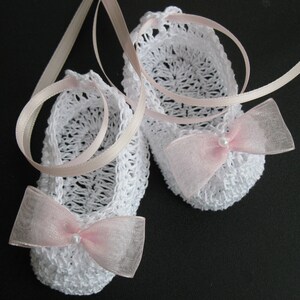 May include: A pair of white crochet baby booties with pink ribbon bows and pearl accents. The booties are connected by a light pink ribbon. The openwork design and delicate bows give the booties a charming, handmade appearance.
