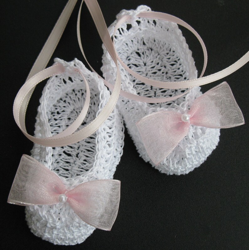 May include: A pair of white crochet baby booties with pink ribbon bows and pearl accents. The booties are connected by a light pink ribbon. The openwork design and delicate bows give the booties a charming, handmade appearance.