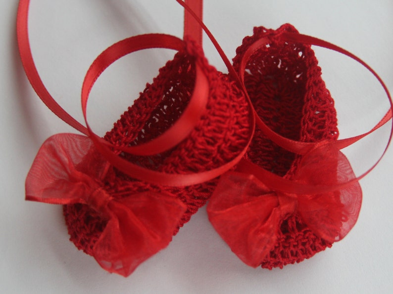 K&ouml;nnte beinhalten: Ein Paar handgefertigte rote Baby-H&auml;kel-Booties mit roten B&auml;ndern und Schleifen aus Chiffon. Die Booties sind aus einem strukturierten Garn gefertigt und f&uuml;r Babys konzipiert.