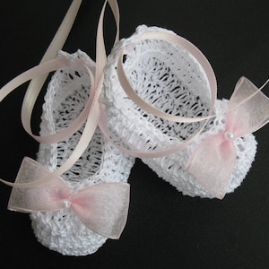 May include: A pair of white crocheted baby booties with pink ribbon ties and delicate pink bows. Each bow is adorned with a small pearl. The booties are suspended against a dark background, showcasing the intricate openwork design.