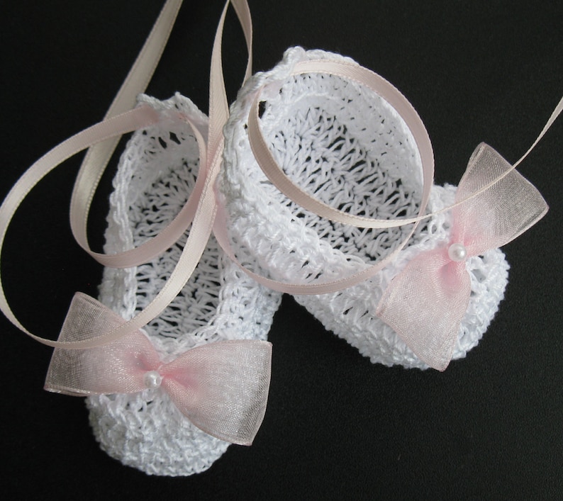 May include: A pair of white crocheted baby booties with pink ribbon ties and delicate pink bows. Each bow is adorned with a small pearl. The booties are suspended against a dark background, showcasing the intricate openwork design.