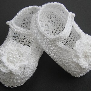Peut inclure: Une paire de chaussons de b&eacute;b&eacute; blancs au crochet. Chaque chaussure pr&eacute;sente une fleur d&eacute;licate avec des perles sur l'orteil et une bride avec un d&eacute;tail de perle. Les chaussures sont de style Mary Jane.