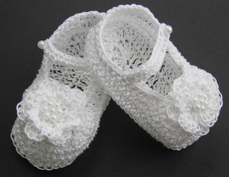 Peut inclure: Une paire de chaussons de b&eacute;b&eacute; blancs au crochet. Chaque chaussure pr&eacute;sente une fleur d&eacute;licate avec des perles sur l'orteil et une bride avec un d&eacute;tail de perle. Les chaussures sont de style Mary Jane.