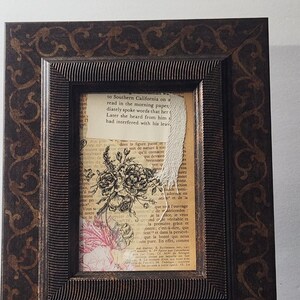 May include: A rectangular picture frame with a dark brown finish and a black scrollwork pattern. The frame holds a collage featuring vintage book pages with English and French text, a black and white floral illustration, and a torn white paper strip.