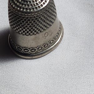 Puede incluir: Un dedal plateado con una parte superior texturizada y una banda decorativa en la base. El dedal est&aacute; sobre un fondo de tela gris claro. El n&uacute;mero "01" es visible.