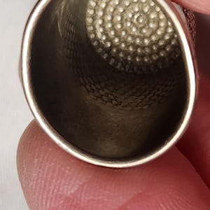 Puede incluir: Primer plano de un dedal plateado. El dedal tiene un interior texturizado y una parte superior circular y con hoyuelos. El dedal est&aacute; sujeto por los dedos de una persona, con un fondo blanco.