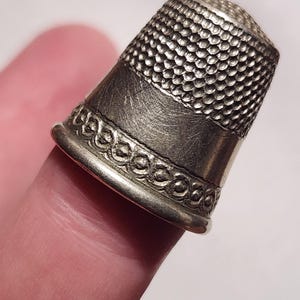 Puede incluir: Primer plano de un dedal plateado. El dedal tiene una parte superior texturizada, una banda lisa y una banda decorativa en la base. El dedal est&aacute; dise&ntilde;ado para coser y hacer manualidades.