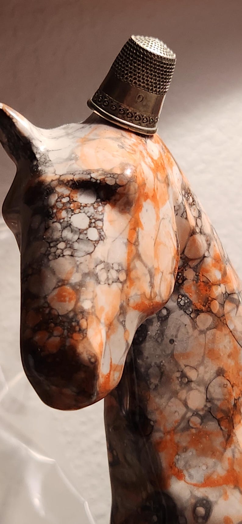 Puede incluir: Escultura de cabeza de caballo de m&aacute;rmol con un dedal plateado en la cabeza. La escultura presenta un patr&oacute;n de remolinos naranjas, blancos y negros. El dedal es plateado con una banda decorativa.