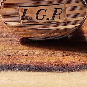 May include: Oval-shaped gold-toned cufflink with a silver-colored clasp. The cufflink features a striped design and the engraved initials "L.G.R." in a rectangular frame. The cufflink is set against a wooden surface.