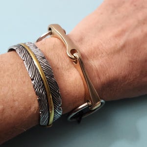 May include: Two bracelets worn together. One is a silver-toned cuff with a textured feather design and a central gold-toned stripe. The other is a gold-toned metal bracelet featuring a central bar connected to two circular rings at the ends.