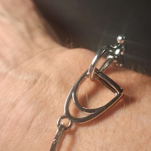 May include: Silver-toned bracelet featuring a unique design with a central, stylized teardrop shape. The bracelet is composed of interconnected links and a decorative element. The jewelry is worn on a wrist.