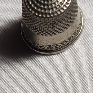 Puede incluir: Un dedal plateado con la parte superior con hoyuelos y una banda decorativa alrededor de la base. La herramienta de costura est&aacute; dise&ntilde;ada para proteger el dedo. El fondo es de tela gris claro.