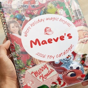 Puede incluir: Un cuaderno de espiral con una portada temática navideña que presenta varios personajes y decoraciones festivas. Una etiqueta circular blanca central muestra el texto en rojo: where holiday magic begins, Maeve's, 2026 toy catalogue, Merry Xmas.