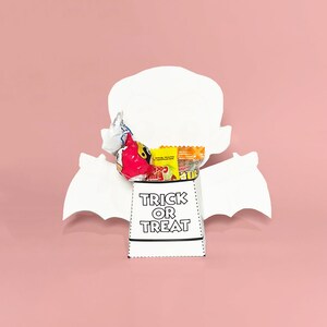 May include: White paper bat-shaped treat bag with the words "Trick or Treat" printed in black. The bag is filled with colorful candies.
