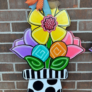 Puede incluir: Un arreglo floral de madera colorido en una maceta de lunares blancos y negros. Las flores son amarillas, moradas, rosas, naranjas y turquesas. La maceta tiene un borde rayado blanco y negro.