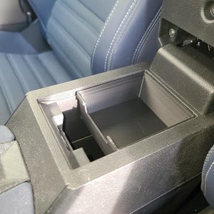 Peut inclure: Console de voiture en plastique noir avec un compartiment de rangement ouvert. La console est intégrée à l'intérieur d'une voiture, avec un siège en cuir bleu visible en arrière-plan. Un bouton rouge "PRESS" est également visible.