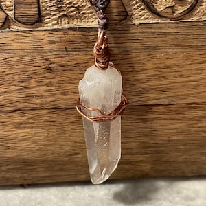 May include: A clear quartz crystal pendant wrapped in copper wire, suspended from a brown braided cord. The crystal is a light, translucent color and has a natural, irregular shape. The pendant hangs against a wooden background.
