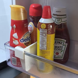 Puede incluir: Un estante de refrigerador con varios condimentos. Incluye una botella roja y amarilla de crema de café International Delight Sweet Cream, jarabe de café, crema batida y una botella de ketchup Heinz.