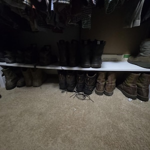 May include: A white wooden shoe shelf filled with various boots and shoes. The shelf is in a closet, with clothing hanging above. The boots are in shades of brown and black, and the carpet is beige.