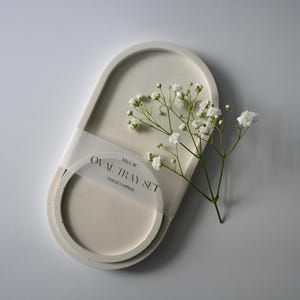 Puede incluir: Un juego de bandejas ovaladas de color blanquecino con una bandeja redonda a juego, envuelto con una banda transparente. El conjunto va acompañado de una ramita de flores blancas. El texto en la banda dice "OVAL TRAY SET" y "Hand crafted".