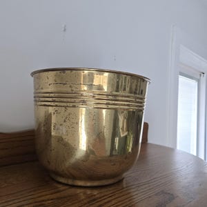 May include: A cylindrical brass planter featuring a polished metallic finish with visible signs of age, including surface patina and minor dark spots. The vessel has a wide, open top and three horizontal ridges encircling the upper rim.