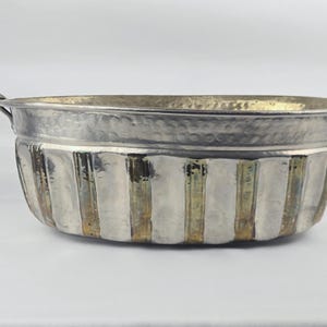May include: An oval-shaped metal basin featuring a hammered texture along the upper rim and vertical fluted panels along the sides. The basin has a metallic silver finish with subtle brass-toned highlights and two small handles on each end.