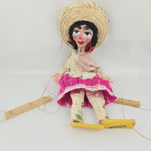 May include: A marionette puppet with black hair, wearing a straw hat, a cream-colored top with floral patterns, and a bright pink skirt with white lace trim. The puppet has painted facial features and is attached to wooden control bars by strings.
